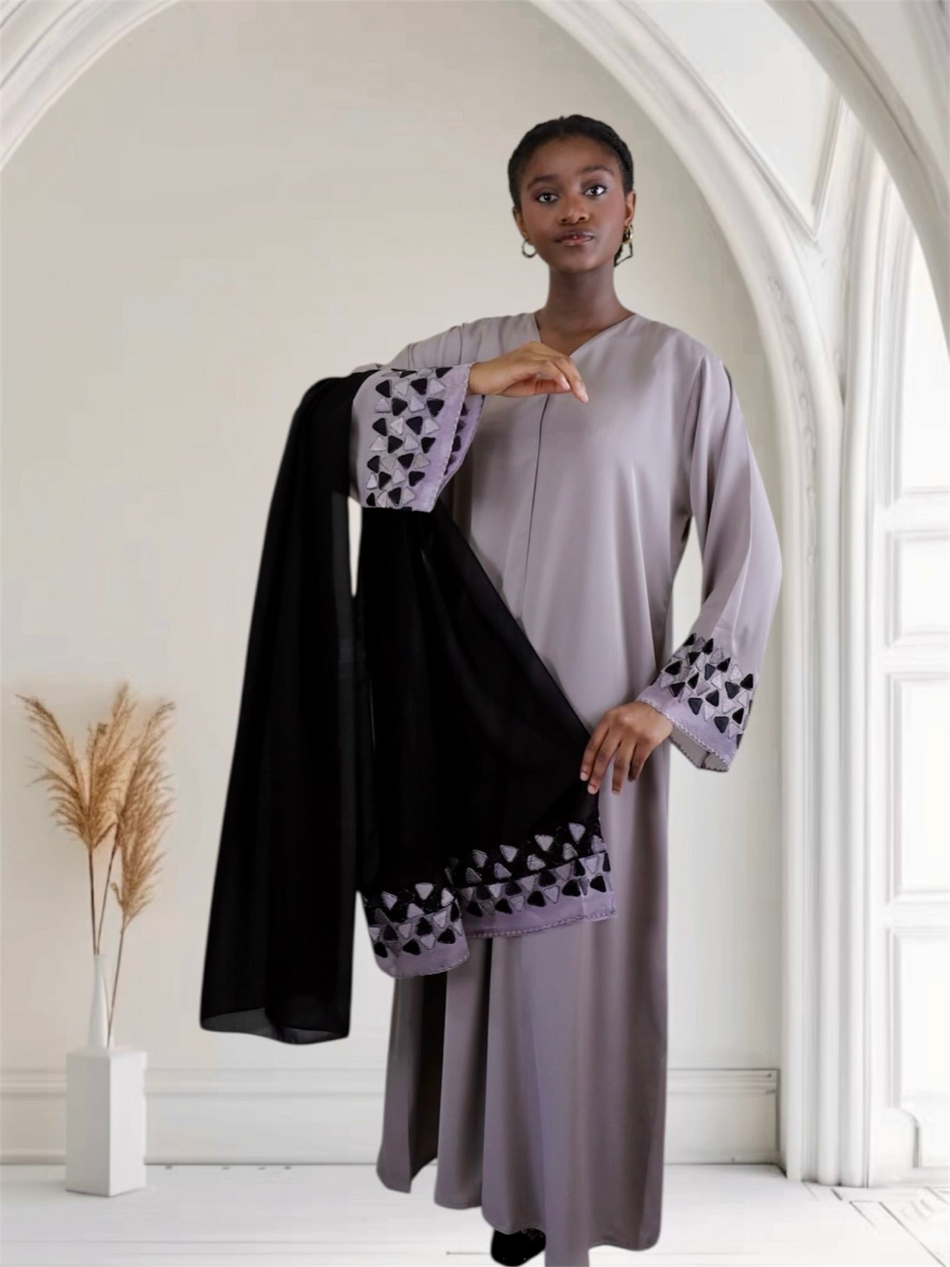 Woman in a light purple outfit with black and white patterned sleeves holding a black scarf in a bright room.