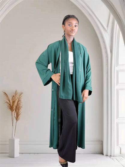 Woman wearing a long green coat in a bright room with white walls and a plant.