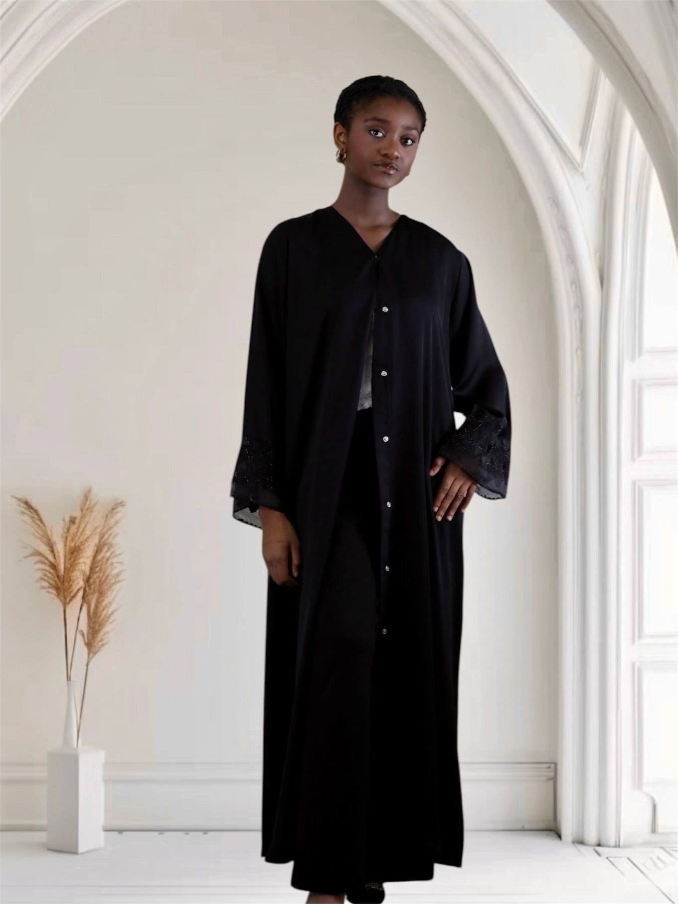 women wearing a black outfit standing in a minimalistic room with white walls and a vase with dried plants.