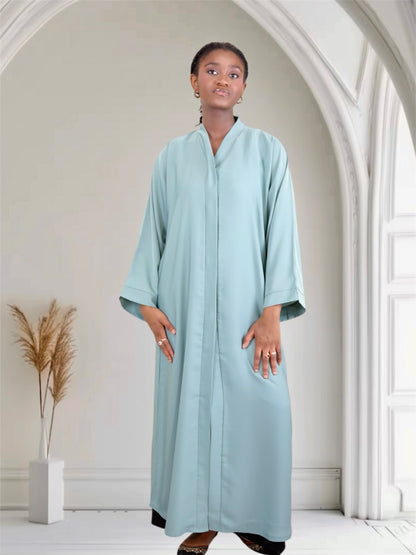 Woman wearing a light blue abaya in a minimalistic room with white walls and a plant.