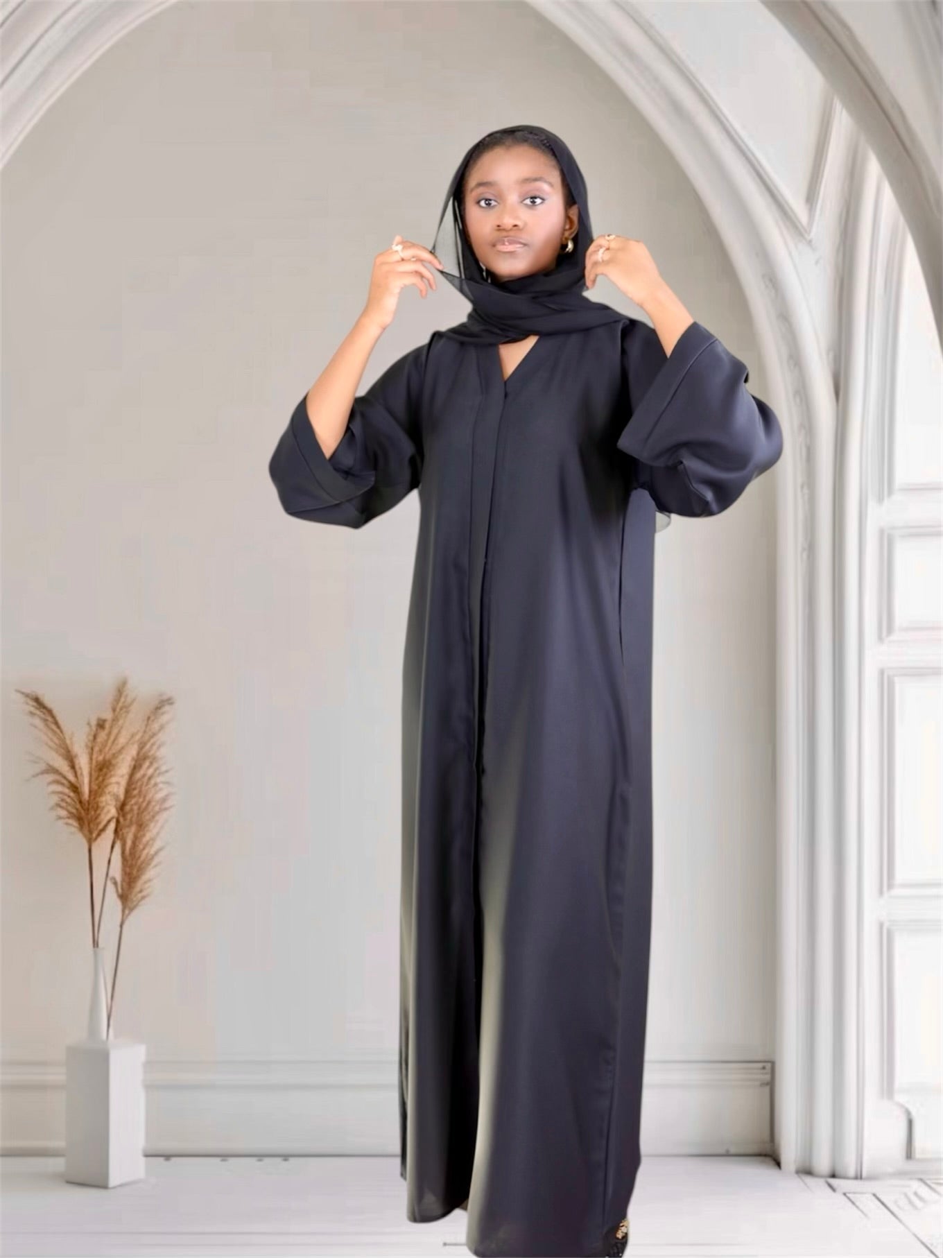 Woman wearing a black abaya in a minimalistic room with white walls and a plant.