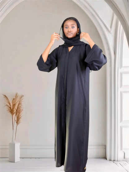 Woman wearing a black abaya in a minimalistic room with white walls and a plant.