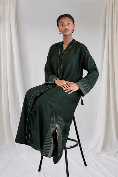 woman wearing a dark green dress sitting on a black stool against a white curtain.