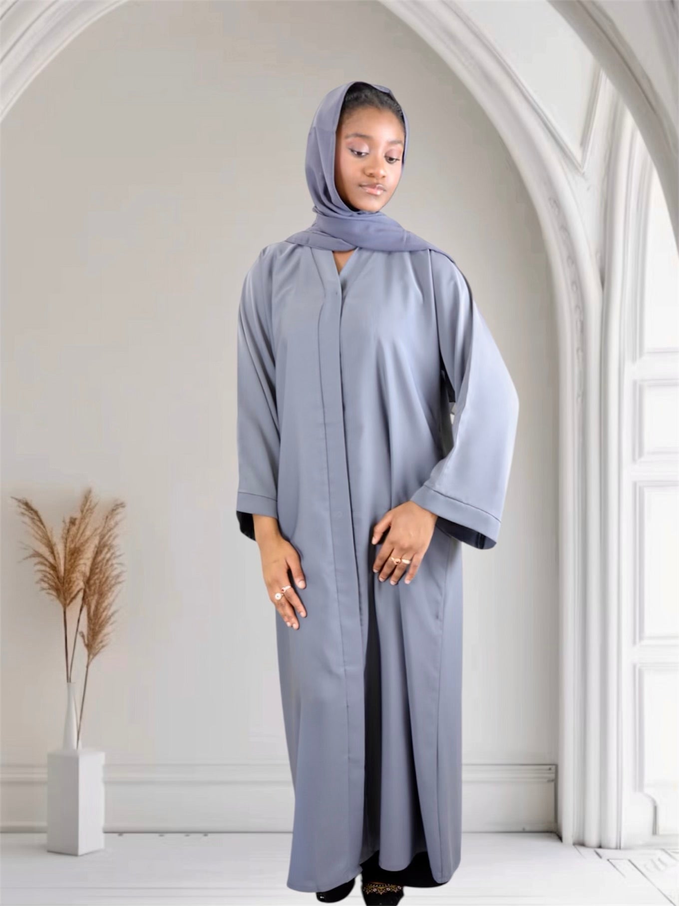 Woman wearing a long, light gray abaya with a matching headscarf in a bright room.